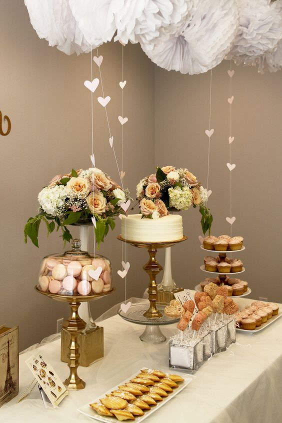 چیدمان میز کیک تولد با ظروف پذیرایی طلایی و گلدان گل و پام پام کاغذی نصب شده در سقف